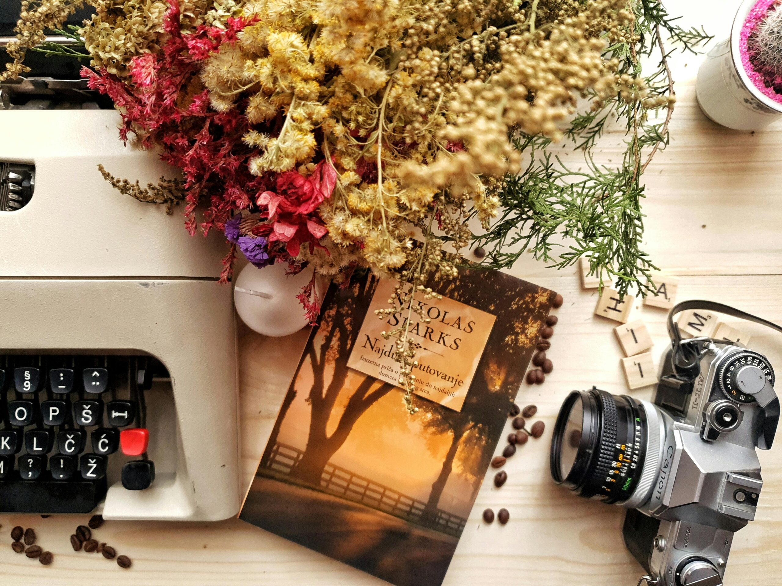 Home Top view of book and photo camera placed on wooden table near vintage typewriter composed with beautiful dried flowers