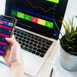 A person using a smartphone app to trade stocks with a laptop displaying market data.