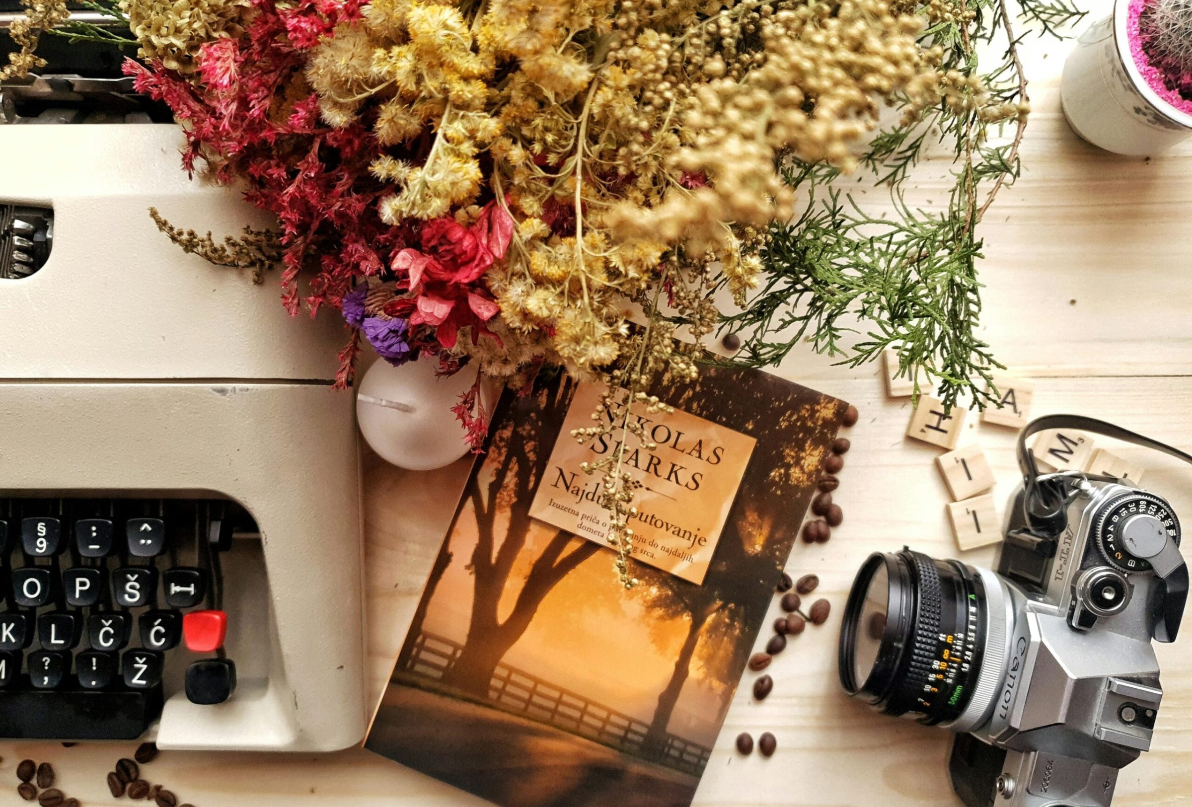 Top view of book and photo camera placed on wooden table near vintage typewriter composed with beautiful dried flowers